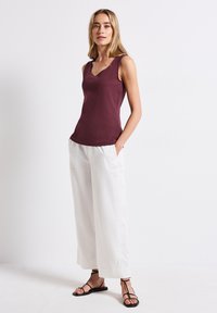 Bordeauxfarbenes ärmelloses Oberteil mit V-Ausschnitt, kombiniert mit weißen weit geschnittenen Hosen. Der Look wird mit schwarzen flachen Sandalen abgerundet.