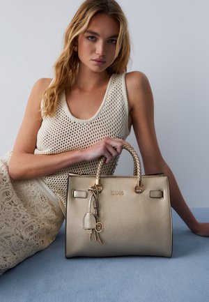 Mlada ženska v bež pleteni obleki drži zlato teksturirano torbico s tkanimi ročaji in pripetimi obeski v obliki ključev, sedi na modri podlagi.