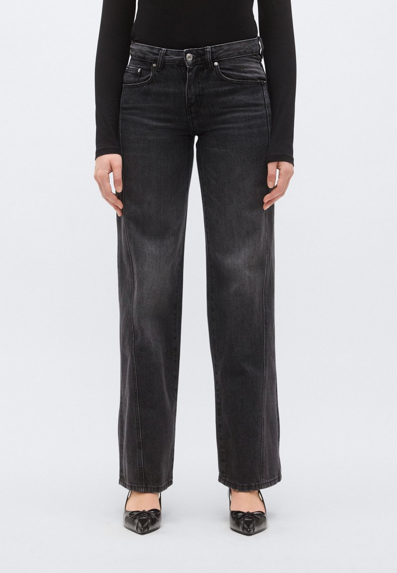 Fekete, széles szárú farmer enyhén kifakult hatással, ötlovas zseb kialakítással és finom steppeléssel. Fekete cipőkkel párosítva, melyeken masni díszítés látható.