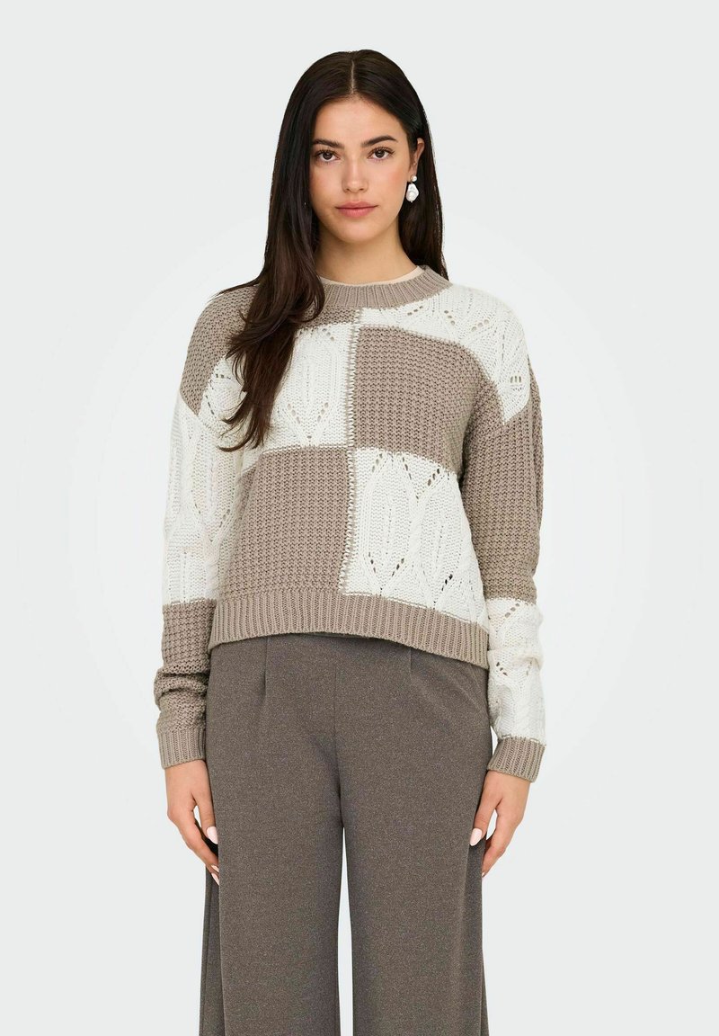 Maglione lavorato a maglia caratterizzato da un design a patchwork in beige e crema, con motivi testurizzati e orlo a costine. Abbinato a pantaloni marroni larghi.