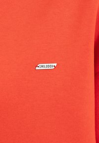 T-shirt a maniche corte in cotone arancione brillante con una texture liscia, dotato di una piccola etichetta in tessuto grigia con "SMILODOX" in testo nero.