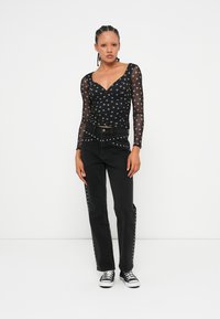 Transparente schwarze Bluse mit langen Ärmeln und silbernen Punkten, kombiniert mit hochtaillierten schwarzen Jeans, die silberne Nietenakzente an den Seiten aufweisen. Schwarze Sneaker.