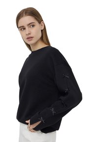Czarny sweatshirt z długimi rękawami, ozdobionymi czarnymi wstążkami przy mankietach. Sweatshirt ma luźny krój i okrągły dekolt.
