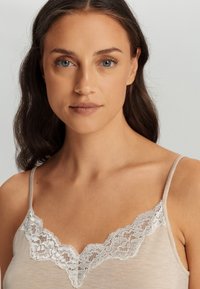 Zachte beige cami met delicate witte kantafwerking langs de v-hals en verstelbare spaghettiriemen. Lichtgewicht stof, gladde textuur.