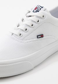 Baskets en toile blanche avec six œillets, lacets plats blancs, et un logo brodé bleu et rouge sur le côté. Semelle en caoutchouc avec surface texturée pour une meilleure adhérence.