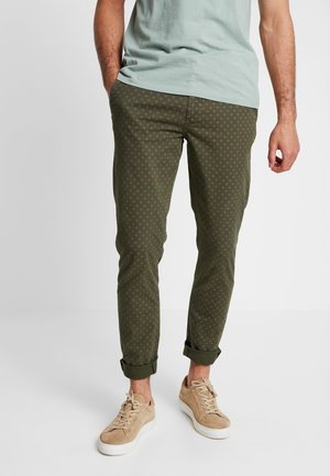 Mand iført op rullede olivengrønne mønstrede bukser, lysegrøn t-shirt og beige sneakers, stående med den ene hånd i lommen.