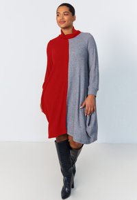 Robe en tricot arborant un design en blocs de couleur rouge et gris, un col montant haut, des manches longues et une poche latérale. Portée avec des bottes noires à hauteur de genoux.