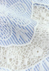 Tissu rayé bleu et blanc avec broderie florale blanche et détails complexes en dentelle sur une surface textile douce et texturée.