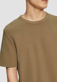 Oliwkowa koszulka z krótkim rękawem z teksturowanej tkaniny. Posiada okrągły dekolt i luźny krój z widocznymi detalami szwów.