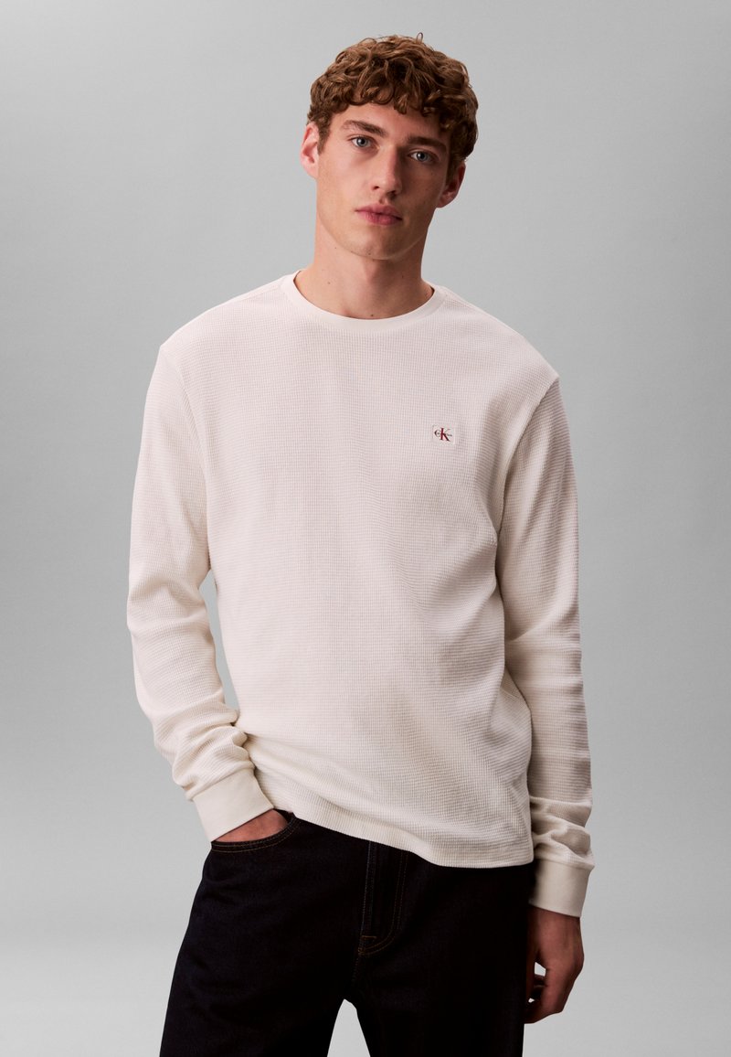 Langærmet cremefarvet termotrøje med tekstureret stof, rund halsudskæring og lille rødt logo på brystet, kombineret med mørke jeans.