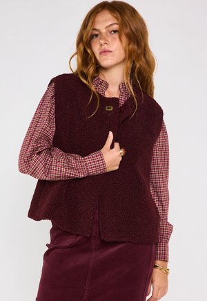 Gilet in shearling bordeaux con orlo arrotondato, chiusura a un bottone, indossato sopra una camicia a quadri rossi e una gonna in velluto a coste coordinata.