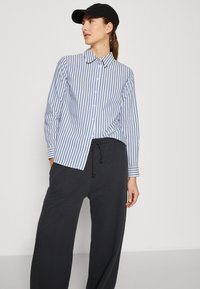Camicia button-up a maniche lunghe con righe verticali blu e bianche, abbinata a pantaloni della tuta neri larghi. Presenta un colletto classico e dettagli sui polsini.