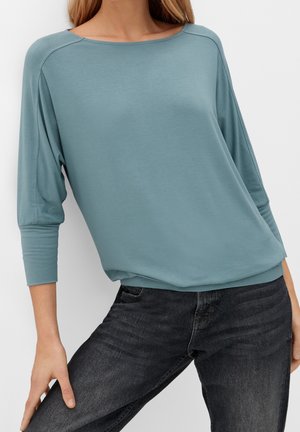 Vrouw met een loszittend tealkleurig shirt met lange mouwen en een boothals, gecombineerd met zwart verweerd spijkerbroek, poserend met haar hand op haar dij tegen een witte achtergrond.