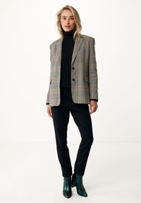 Blazer sartorial a scacchi beige e nero, maglione nero a collo alto, pantaloni neri su misura e stivaletti verde lucido a caviglia.