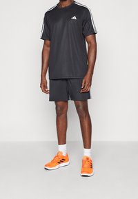 Černé sportovní tričko s bílými pruhy a logem, odpovídající černé šortky a jasně oranžové boty s černými akcenty, nošené s bílými ponožkami.