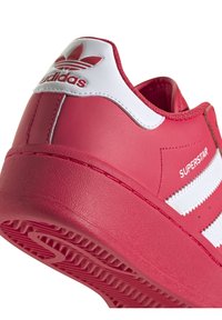 Röd Adidas Superstar-sneaker med vita ränder, texten "SUPERSTAR" på sidan och Adidas trefoil-logotyp på hälen.