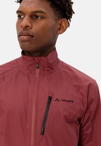 Rote leichte Jacke mit hohem Kragen und einem Frontreißverschluss. Verfügt über eine schwarze Reißverschlusstasche und ein Logo auf der linken Brustseite. Glatte Textur.