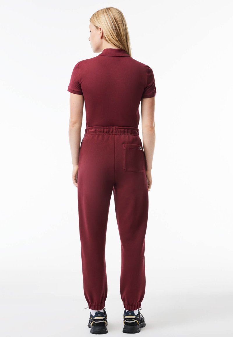 Completo bordeaux con maniche corte, collo a camicia, cintura elastica con tasca e caviglie risvoltate. Finitura in tessuto testurizzato.