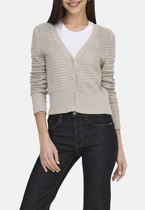 Gilet tricoté beige avec un col en V, fermeture à boutons et motif texturé. Porté sur un haut blanc avec un jean en denim foncé.
