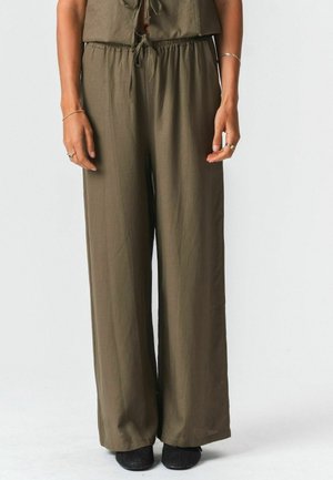 Kvinde iført olivengrønne vidde benbukser med snøre i taljen og sorte teksturerede slip-on sko, stående mod en ensfarvet baggrund.