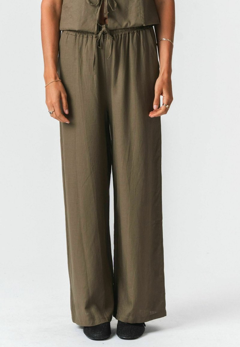 Femme portant un pantalon large vert olive à taille élastique avec cordon de serrage et des chaussures plates noires texturées, debout devant un fond uni.