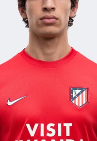 Jeune homme portant un maillot de football rouge avec l'écusson de l'Atletico Madrid, le logo Nike et un texte partiel du sponsor "Visit Rwanda".