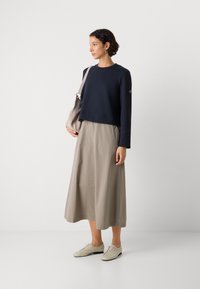 Mujer de pie, vistiendo una camiseta de manga larga azul marino y una falda midi beige, sosteniendo un bolso de hombro gris, con zapatos blancos con cordones, sobre un fondo blanco.
