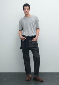 T-shirt grigio a maniche corte, jeans neri con risvolti e mocassini marroni. Un maglione blu è annodato in vita. Ambientazione minimalista.