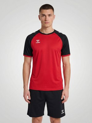 Jonge man staand, gekleed in een rood-zwart sportshirt en zwarte short met zichtbare witte hummel-logo's op borst en short.