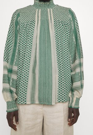 Langærmet bluse med grønne og beige geometriske mønstre, høj smock-krave og matchende smock-manchetter, kombineret med lyse beige bukser.