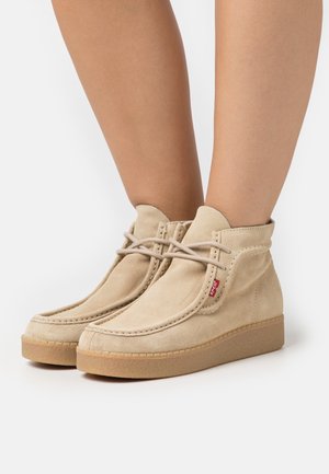Beige Wildleder-Stiefeletten mit Gummisohlen und Schnürdetail, getragen an nackten Beinen vor weißem Hintergrund.