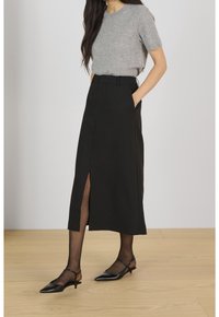 Jupe midi noire avec une fente avant, assortie à un haut tricot à manches courtes gris. Complétée par des collants noirs transparents et des chaussures noires à talons pointus.