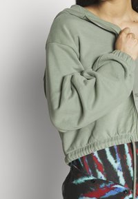 Grøn oversized hoodie med lynlås, samlet ærmer og ribbet kant. Modellen har den på over mønstrede sorte og farverige bukser.