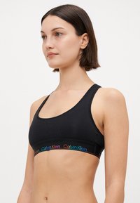 Svart bomulls-bralette med scoop neckline, tynne stropper og en flerfarget Calvin Klein-logo i midjen. Myk tekstur, sømløst design.