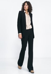 Kobieta z kręconymi włosami, ubrana w czarny garnitur, beżową bluzkę i beżowe buty, stojąca na białym tle, patrząca w bok.