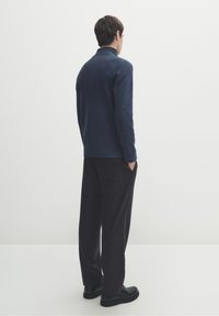 Pull-over en maille bleu foncé avec col côtelé, associé à un pantalon ample gris charbon. Le modèle se tient de dos, les mains dans les poches.