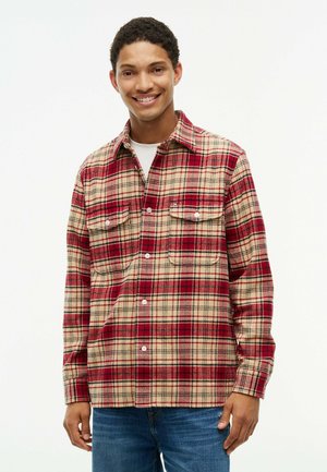 Ung mand iført en rød og beige ternet skjorte over en hvid t-shirt, stående og smilende mod en ensfarvet baggrund.