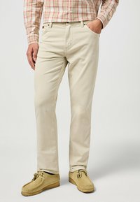 Los pantalones de algodón beige presentan un diseño de pierna recta, bolsillos frontales y una textura suave, combinados con zapatos estilo mocasín de color beige claro.