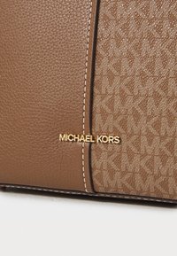 Ruskea nahkainen käsilaukku, jossa on vaaleanruskeat yksityiskohdat, ja jossa on teksturoitu kangasosa sekä kultainen "Michael Kors"-logo. Korostettu valkoisella ompelulla.