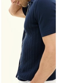 Navyblauwe gebreide polo met korte mouwen, met een textuurpatroon, witte knoopsluiting en contrasterende afwerkingsdetails.