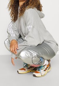 Haut à capuche gris surdimensionné avec des rayures blanches sur les manches, leggings argentés brillants avec des lignes horizontales, et baskets de sport multicolores.