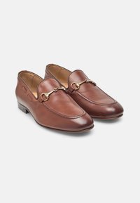 Mocassins en cuir marron avec des garnitures en métal doré, bouts ronds et détails cousus. Texture lisse avec un petit talon et des découpes sur les côtés.