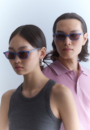 Blaue Sonnenbrille mit rechteckigen Gläsern, kombiniert mit einem ärmellosen grauen Oberteil. Der Hintergrund ist weich und hell unscharf. Das Model trägt ein hellrosa Poloshirt.