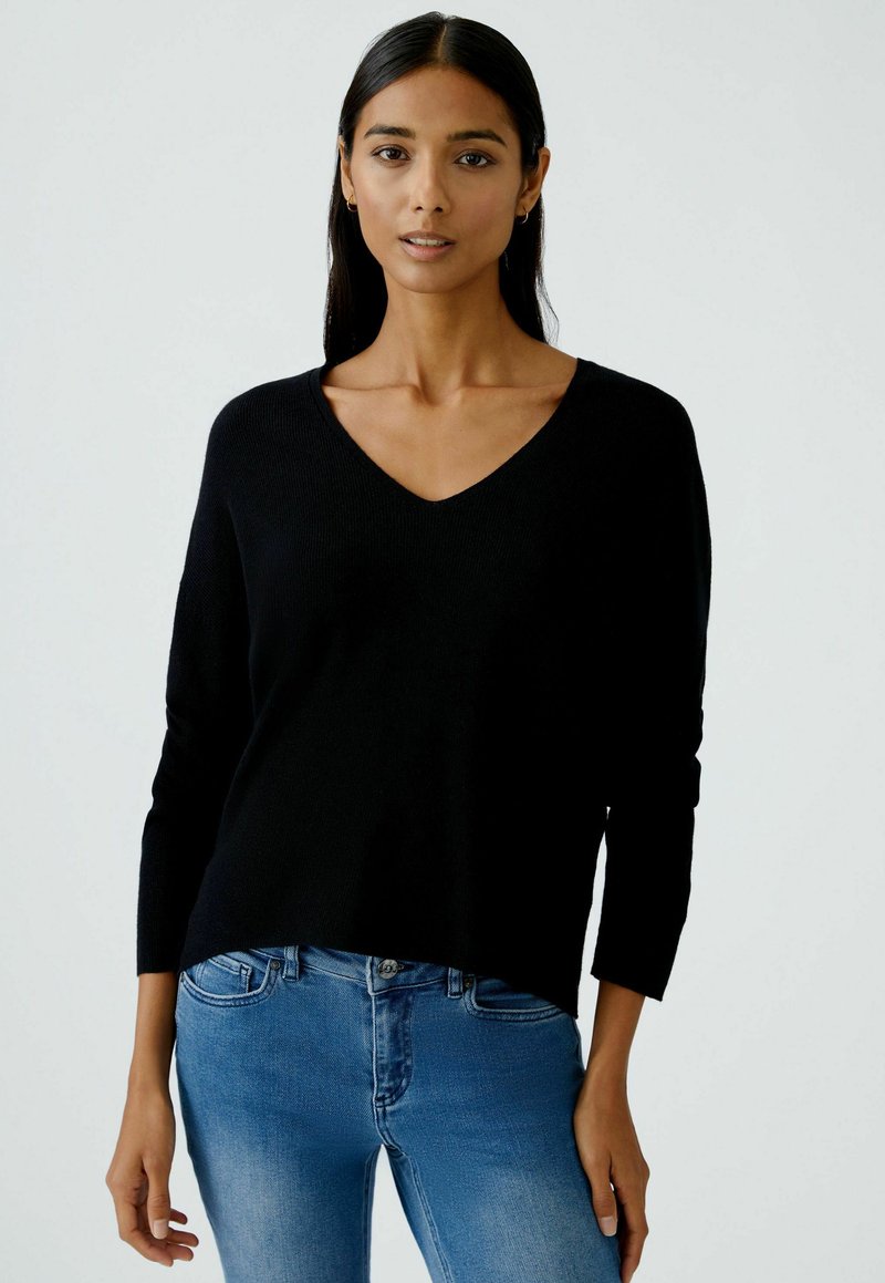 Schwarzer V-Ausschnitt-Pullover mit langen Ärmeln, gerippter Textur und lässiger Passform, kombiniert mit hellblauen Jeans, vor einem neutralen Hintergrund präsentiert.