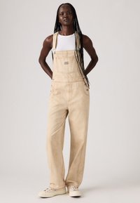 Beige denim overalls with wide legs, adjustable straps, and front pocket detail; worn over a fitted white tank top. Casual footwear complements.