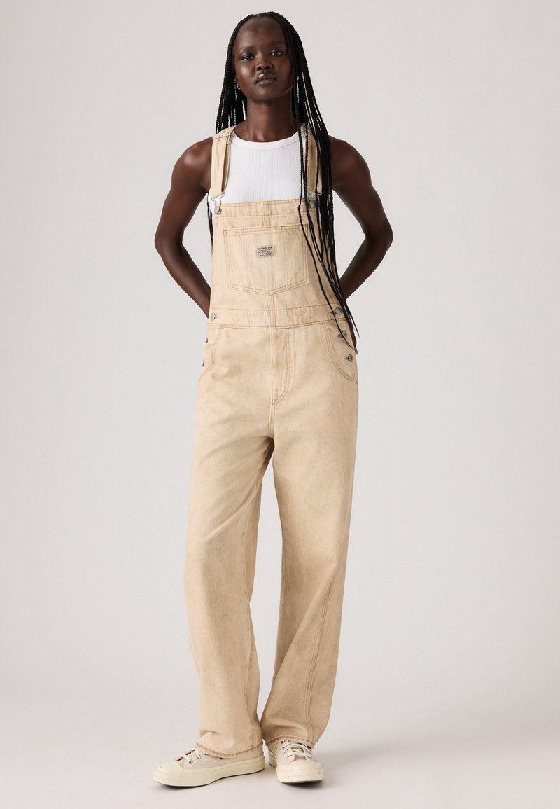 Beige denim overalls with wide legs, adjustable straps, and front pocket detail; worn over a fitted white tank top. Casual footwear complements.
