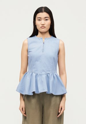 Νεαρή γυναίκα που φοράει αμάνικο, ανοιχτό μπλε peplum τοπ με φερμουάρ μπροστά και φαρδιά ελαιέρια παντελόνια, στέκεται μπροστά σε απλό φόντο.