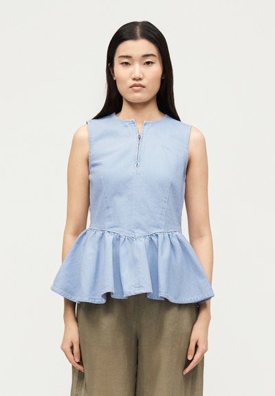Jeune femme portant un top peplum bleu clair sans manches avec une fermeture éclair à l'avant et un pantalon ample couleur olive, debout sur un fond uni.