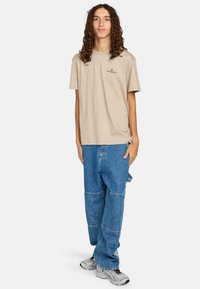 Camiseta de algodón beige con mangas cortas y un pequeño logo, combinada con pantalones de mezclilla azul holgados que presentan detalles de parches y un corte relajado.