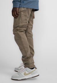 Cargo pants in lichtbruin katoen met meerdere zijzakken, gecombineerd met grijze en crèmekleurige sneakers met gouden accenten.